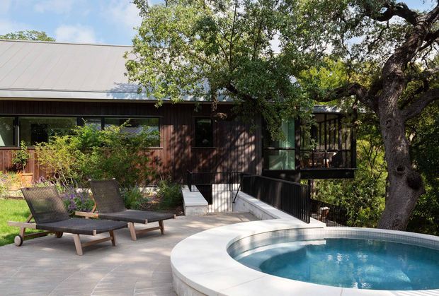 Exterior view of a contemporary single-storey home integrated into a leafy wooded setting. The dark timber-clad facade has extensive glazing, a light standing-seam metal roof, and a raised deck or balcony. In the foreground, a paved terrace with outdoor loungers surrounds a curved, partially visible swimming pool.