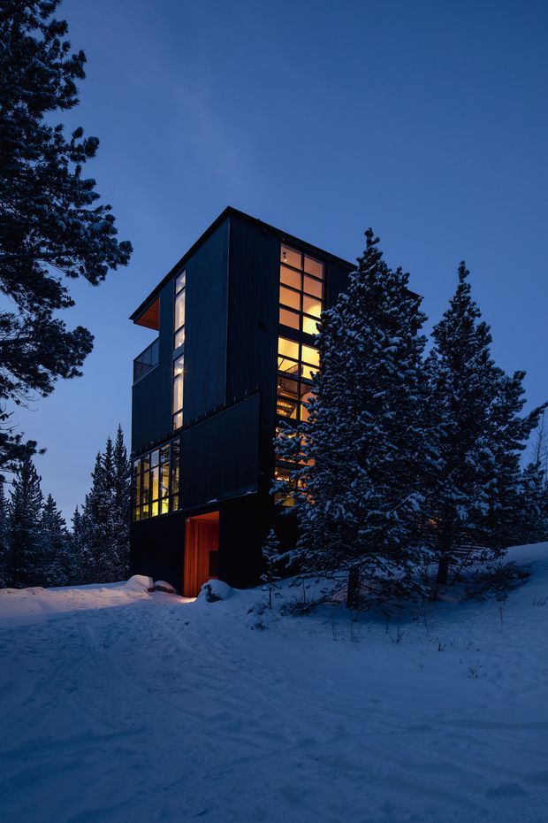A tall, narrow multi-storey modern house set among snow-covered evergreen trees at dusk. The dark vertical facade has large floor-to-ceiling glazed sections glowing warmly from within, with projecting volumes, balconies, and a sheltered illuminated entrance. The surrounding alpine woodland and deep snow create a dramatic winter setting.