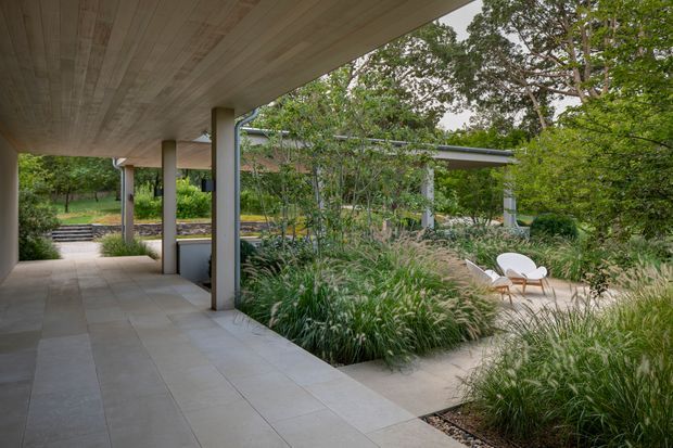 A covered outdoor walkway or terrace with a broad timber-lined soffit, slender structural columns, and large pale stone or concrete-look floor tiles. The space opens onto a landscaped courtyard filled with ornamental grasses, shrubs, and mature trees, with a small outdoor seating area visible beyond. The architecture is restrained and contemporary, with strong indoor-outdoor connections and a calm, garden-focused atmosphere.