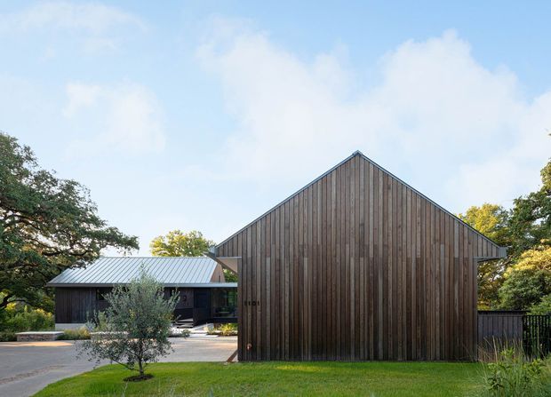 Exterior view of a contemporary residential property featuring a prominent gabled structure clad in vertical dark timber boards, with a low adjoining wing topped by a light grey metal roof. The house is set within mature landscaped greenery and a lawn, with a paved forecourt and glimpses of glazing and entry areas.