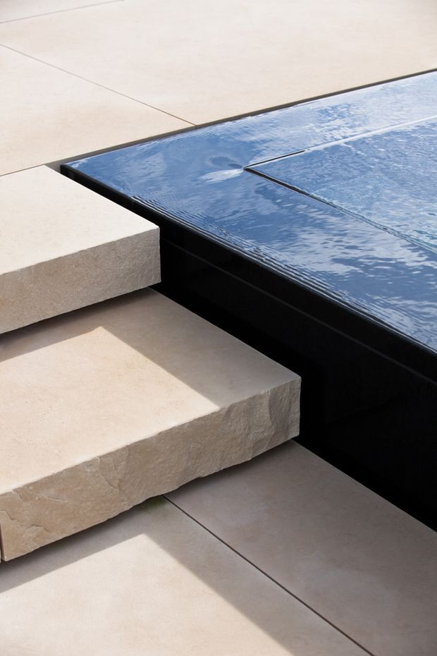 Close-up architectural detail of broad pale stone steps meeting the edge of a dark-lined swimming pool. Large beige paving slabs surround rippling blue water, with crisp geometric junctions and strong sunlight creating a modern, refined outdoor setting.