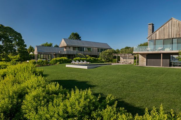 Wide exterior view of a contemporary multi-building residential property set within expansive landscaped lawns. The buildings feature steep gabled forms, dark standing-seam metal roofs, vertical timber cladding, large glazed openings, balconies, and a central in-ground swimming pool. Mature planting and hedges surround the home.