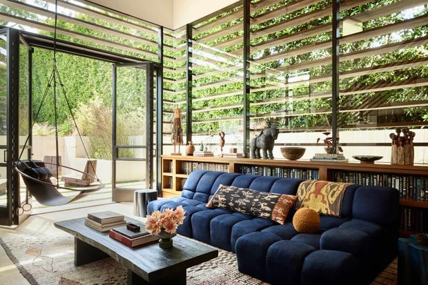 A sophisticated living room with a deep navy tufted sectional sofa, dark rectangular coffee table, patterned cushions, and a large textured rug. Floor-to-ceiling glazing and open glass doors connect the room to a lush garden, while horizontal timber screening provides filtered light and privacy. Built-in timber shelving filled with books and decorative objects runs along the rear wall, and a suspended swing chair adds a distinctive feature.