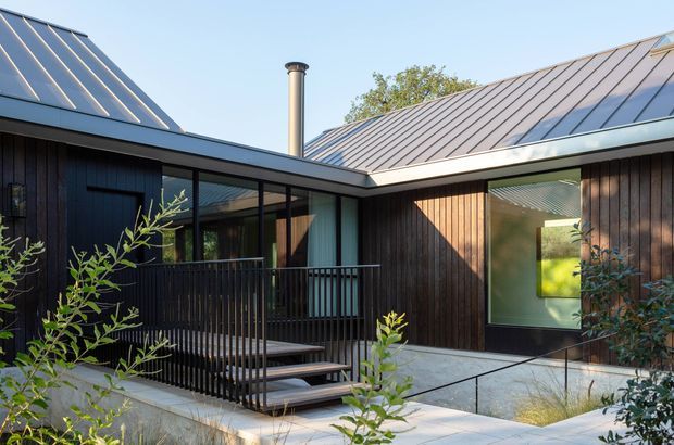 Exterior view of a contemporary single-storey home arranged around a sheltered courtyard. The building has dark vertical timber cladding, large black-framed glazed openings, standing-seam metal roofs with multiple pitched planes, a covered deck with black metal balustrades, and pale concrete paving. Native-style planting partially frames the foreground.