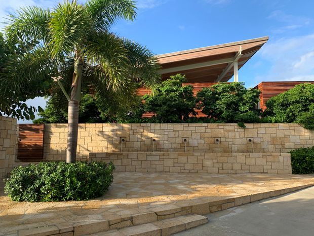 Exterior view of a contemporary single-storey residence behind a low tan stone boundary wall, with warm horizontal timber cladding, a broad sloping roof canopy, landscaped tropical planting, and paved stepped frontage.
