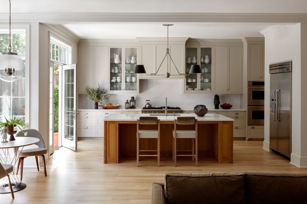 A bright, spacious traditional-style kitchen viewed from an adjoining living or dining area. The room features a large central timber island with seating for three, pale painted cabinetry, glass-front display cupboards, a light stone benchtop, integrated stainless-steel appliances, timber flooring, and large glazed doors and windows. A simple geometric pendant hangs above the island, while a small dining area and sofa edge are visible in the foreground.