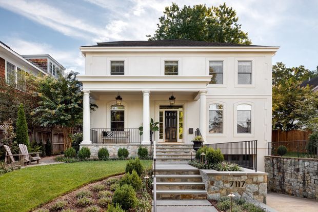 Front exterior view of a refined two-storey residence with a symmetrical cream-painted facade, shallow hipped roof, classical porch supported by round columns, decorative window surrounds, dark entry door, front steps, landscaped garden, and stone retaining walls.
