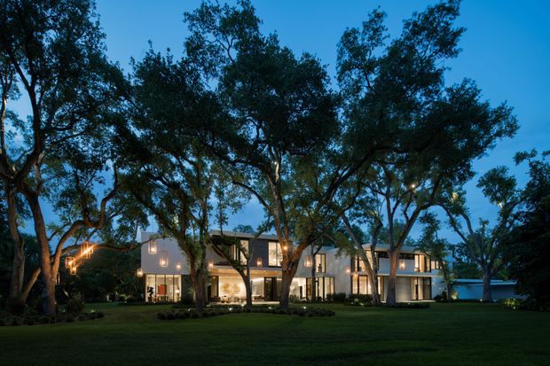 Twilight exterior view of a large contemporary multi-wing residence set within a mature, tree-filled landscaped site. The building has pale rendered or masonry walls, extensive floor-to-ceiling glazing, warm interior lighting, and a low-pitched roofline, with a broad lawn and mature canopy trees in the foreground.