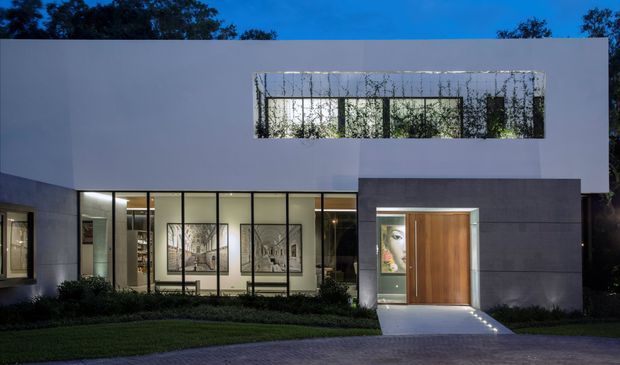 Night-time exterior view of a modern two-storey residence with a clean rectilinear form, smooth white upper walls, dark grey lower cladding, extensive floor-to-ceiling glazing, and a large timber pivot entry door. A long upper-level window is screened by climbing vegetation, while illuminated interiors are visible through the ground-floor glazing. The house is set behind landscaped planting and a paved driveway.