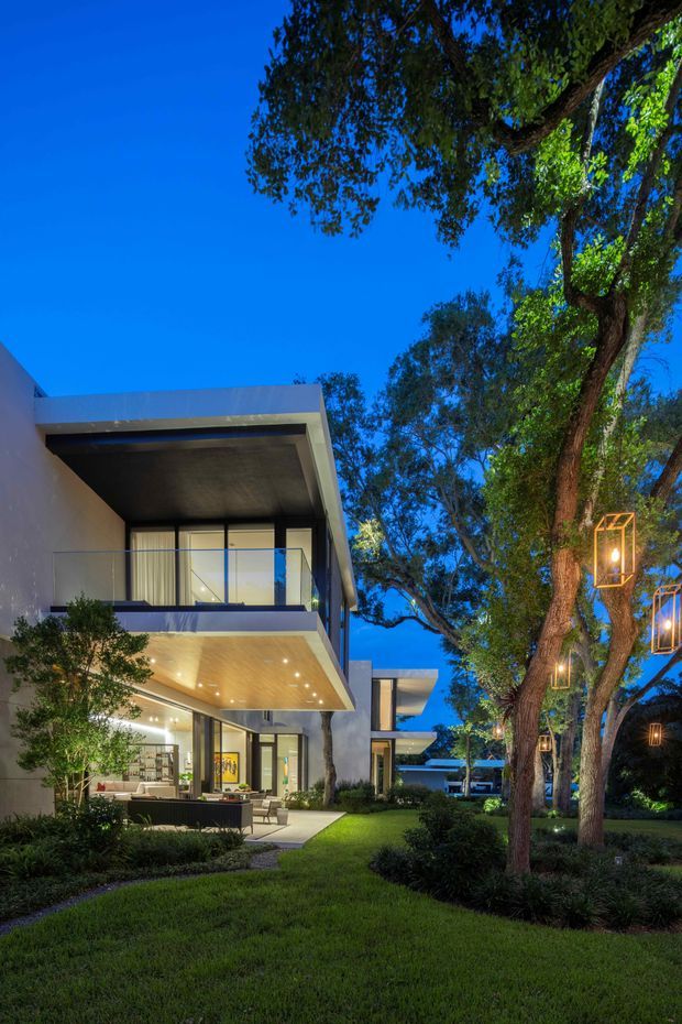 Twilight exterior view of a contemporary multi-level residence set within a landscaped garden. The home features crisp flat rooflines, projecting cantilevered balconies and roof overhangs, expansive glass doors and windows, slim dark frames, and warm timber soffits. Mature trees surround the lawn and are decorated with suspended geometric lanterns, creating a welcoming illuminated outdoor setting.