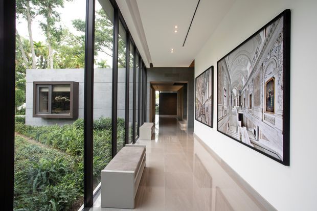 A long, refined interior gallery corridor with full-height black-framed glazing along one side, a lush garden visible outside, polished pale stone flooring, crisp white walls, and minimalist recessed linear lighting. Large framed architectural artworks line the opposite wall, while a simple built-in bench runs beside the windows. The corridor leads toward a darker timber-toned transition space at the far end.