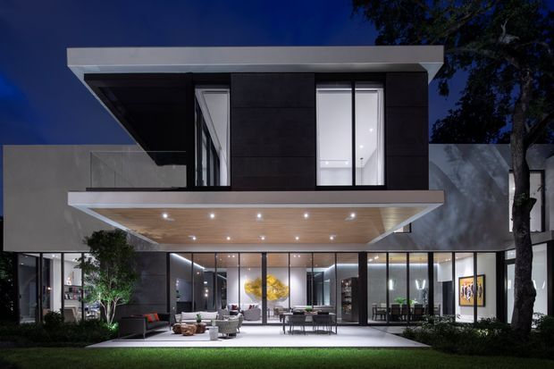 Night-time exterior view of a contemporary two-storey residence with a strong flat-roofed composition, dark upper-level cladding, expansive floor-to-ceiling glazing, and a large cantilevered covered terrace or entry canopy. Warm recessed lighting under the canopy illuminates the outdoor entertaining area and visible open-plan interiors, with landscaped planting and lawn framing the foreground.