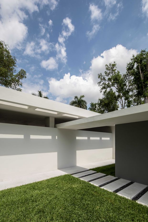 Minimalist modern exterior courtyard with crisp white rendered walls, overlapping flat roof planes, narrow clerestory openings, and a concrete stepping-stone path set into a manicured lawn. Tropical trees and a bright blue sky are visible above the low-slung architecture.