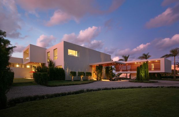 A twilight exterior view of a large contemporary residence with interconnected flat-roofed cubic volumes, smooth light-coloured walls, expansive glazing, and a covered entrance or carport linking the wings. The landscaped frontage includes clipped hedges, tall shrubs, lawn, and a gravel driveway. Warm interior and exterior lighting contrasts with the blue and pink evening sky.