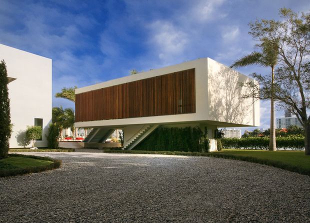 Exterior view of a modern elevated rectangular building with crisp white rendered walls, a large vertical timber screening panel across the upper facade, and open supports beneath. A broad gravel driveway and landscaped gardens surround the structure, with palms and other trees visible against a blue sky.
