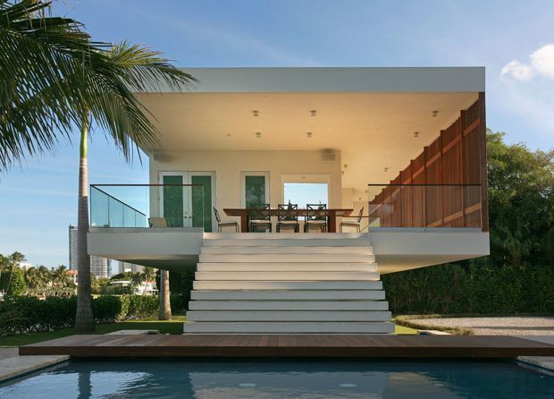 Front exterior view of a contemporary elevated pavilion-like house overlooking a swimming pool. The open upper level features a covered outdoor dining and living area, broad white steps, frameless glass balustrades, and a warm timber privacy screen on the right. Palm landscaping and a flat roof reinforce the tropical modern setting.