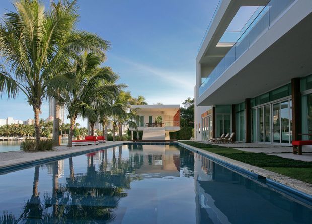 A contemporary multi-level residence arranged around a long rectangular swimming pool. The scene features white rendered volumes, extensive floor-to-ceiling glazing, glass balcony balustrades, broad cantilevered roof or balcony slabs, and a landscaped pool terrace with palm trees and outdoor seating. Bright blue sky and water create a clean, resort-like atmosphere.