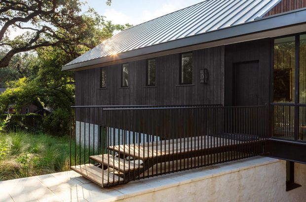 Exterior view of a contemporary raised house set among mature trees and dense landscaping. The building has dark vertical timber cladding, a standing-seam metal gable roof, large dark-framed glazing, and a covered elevated terrace or balcony with black metal balustrades. Pale rendered or concrete masonry supports form the lower level, with stairs leading down to the garden.