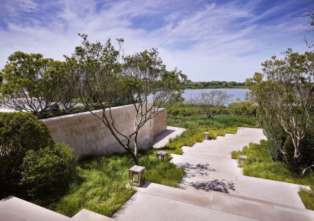 A landscaped outdoor walkway descends through dense ornamental grasses and carefully pruned trees toward a calm body of water. Broad pale concrete steps and paths are paired with low rectangular lanterns and a substantial warm-toned masonry privacy wall, creating a serene contemporary garden setting.