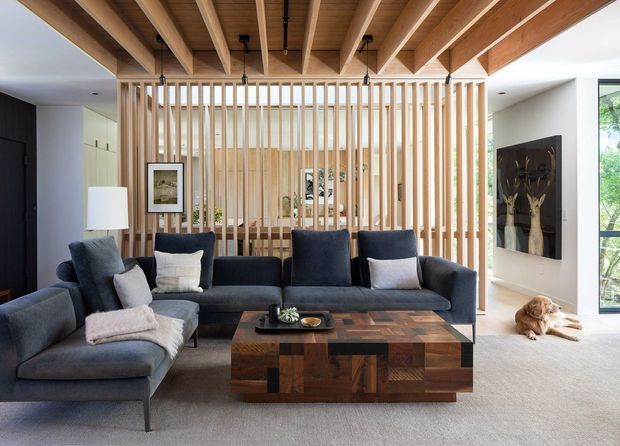 A refined contemporary living room with a large charcoal sectional sofa arranged around a substantial patchwork timber coffee table. A full-height vertical timber slat screen divides the seating area from the space beyond, while exposed timber ceiling beams add warmth and architectural character. The room includes dark feature walls, pale carpet, soft neutral cushions, framed artwork, and generous glazing to the right.