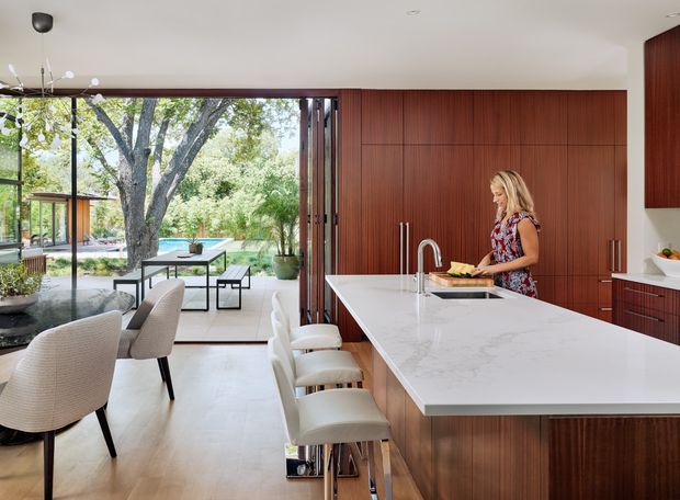 A refined open-plan kitchen and dining area with a large white stone island and integrated seating. Warm vertically grained timber cabinetry forms a full-height feature wall, while floor-to-ceiling glazed doors open to a landscaped outdoor setting and pool area. Neutral upholstered dining chairs and bar stools create a calm, contemporary interior with strong indoor-outdoor connection.