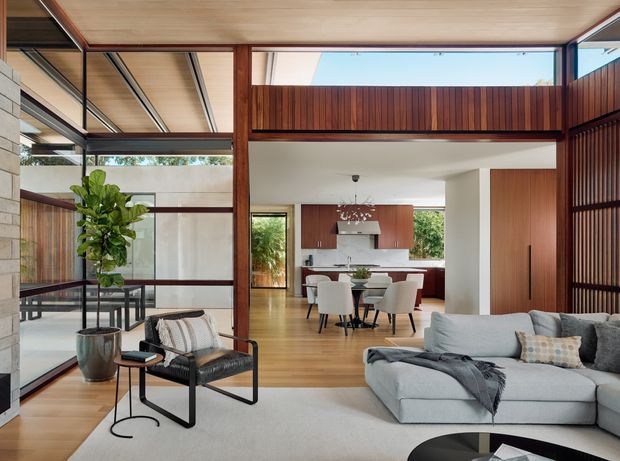A refined open-plan living room looking through to a dining area and kitchen. The space features extensive floor-to-ceiling glazing, warm timber framing and ceiling panels, dark framed windows, timber flooring, a pale sectional sofa, and a black-framed lounge chair. The kitchen and dining zone are visible beyond the living area, with a strong indoor-outdoor connection and abundant natural light.