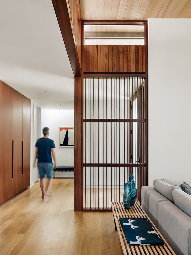 A refined contemporary interior showing a timber-lined hallway and living area. A tall dark timber-framed screen with vertical slats separates the circulation zone from the lounge, extending to a light timber ceiling and clerestory opening. Pale timber flooring, full-height timber cabinetry, a neutral sectional sofa, and a slatted timber bench create a warm, minimalist atmosphere.