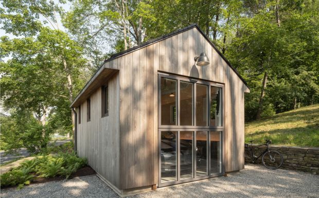 Exterior view of a compact, single-storey gable-roof building clad in vertical natural timber boards. The front facade has large multi-pane glazed doors or windows with dark frames, a wall-mounted exterior light, and a gravel approach. The structure sits among mature woodland with a low stone retaining wall and bicycle visible nearby.