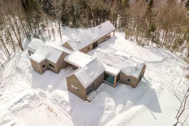 Aerial view of a low-rise timber-clad house nestled in a snow-covered woodland setting. The irregular, multi-winged form has several pitched roof sections heavily blanketed with snow, along with glazed connections and rectangular windows. Snow-covered paths and surrounding forest emphasize its secluded alpine environment.