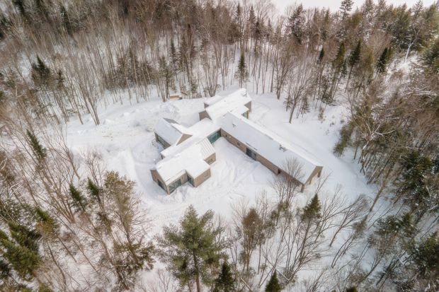 Aerial view of a low-rise, angular residential building nestled in a dense snow-covered forest. The building has multiple interconnected wings with mostly flat or low-pitched snow-covered roofs, pale exterior walls, and a small section of green-toned roofing. Curving paths and driveway-like clearings are visible through the surrounding winter woodland.