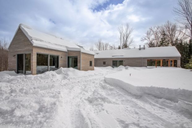 A low, sprawling contemporary house photographed in deep winter, surrounded by heavy snow and leafless trees. The building has pale horizontal wood cladding, simple gabled and low-pitched roof forms covered in snow, and extensive dark-framed glazing, including a glazed projecting wing and a covered timber-framed opening. Snow-covered paths and drifts dominate the foreground.