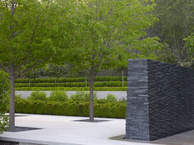 A landscaped contemporary outdoor courtyard or entry area featuring pale stone paving, mature deciduous trees, clipped hedges, and layered garden planting. A long sculptural wall made from dark stacked stone forms a strong architectural feature, with a contrasting lighter stone end section. The scene has clean lines, restrained materials, and a formal, carefully designed garden setting.
