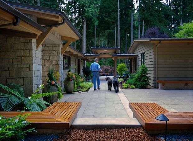 A landscaped residential courtyard walkway between low-rise buildings in a wooded setting. The scene features natural stone wall cladding, timber-framed covered walkways and exposed roof eaves, grey paved flooring, built-in timber lounge benches, abundant planting, and a person walking two dogs. The architecture has a warm, restrained contemporary character integrated into the forested site.
