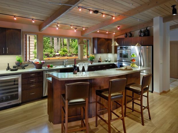 A spacious, warm-toned kitchen with a large central island and seating for four. The room features dark timber cabinetry, pale stone benchtops, stainless-steel appliances, timber flooring, and a timber-lined ceiling with exposed beams and track lighting. Large windows overlook a planted outdoor area, creating a strong connection to nature.