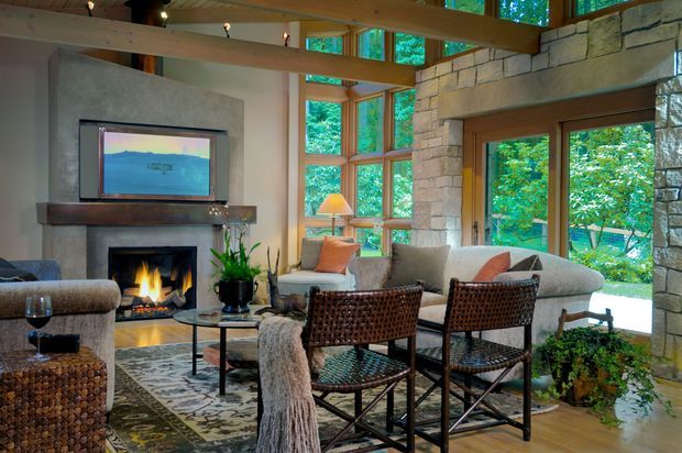 A warm, rustic living room centered around a stone fireplace with a timber mantel and television above. Exposed timber beams, extensive timber-framed glazing, and a large stone-clad wall create a lodge-like connection to the surrounding greenery. The room includes upholstered seating, woven leather chairs, a coffee table, patterned rug, indoor plants, and a lit fire.