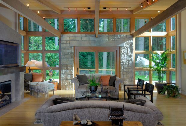 A warm, spacious living room with expansive timber-framed glazing overlooking dense greenery. The room features exposed heavy timber beams, a stone-clad fireplace wall with built-in television, timber flooring, upholstered seating arranged around a glass coffee table, and potted plants. Track lighting and abundant windows create a bright, nature-connected atmosphere.