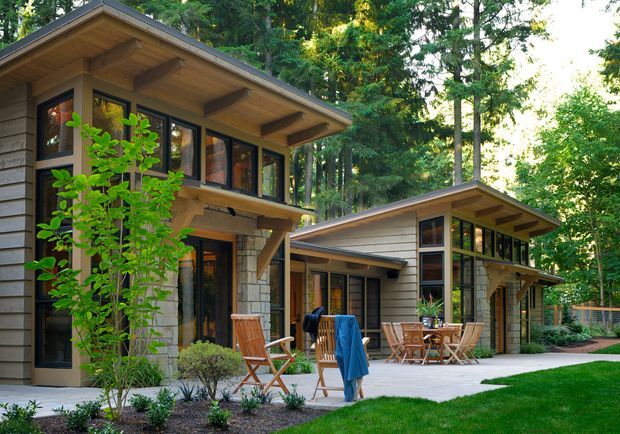 Exterior view of a contemporary two-wing residential house set within a wooded garden. The home features low-pitched gabled roof forms with broad timber soffits and exposed rafters, horizontal light-coloured cladding, dark-framed glazing, stone veneer columns and walls, and covered outdoor dining and seating areas connected by a paved terrace.
