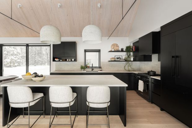 A contemporary kitchen viewed from the dining area, featuring a large island with three upholstered bar stools, matte black cabinetry and appliances, a pale stone benchtop and splashback, light timber flooring, and a striking light timber ceiling with black linear detailing. Large glazed doors/windows bring in a snowy outdoor view, while woven pendant lights and open shelving add warmth.