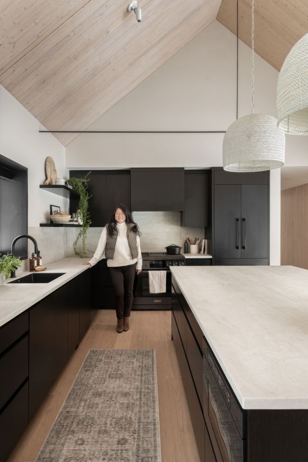 A refined contemporary kitchen with a compact galley-like layout and a large central island. The space features matte black flat-panel cabinetry, pale stone benchtops, a matching stone splashback, light timber flooring, and a dramatic pitched ceiling lined in pale timber. Two woven pendant lights hang above the island, while open shelving, integrated appliances, a sink, cooktop and understated decor complete the warm, modern interior.