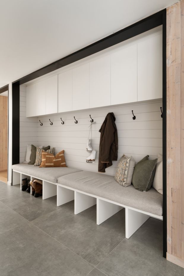 A built-in mudroom or entryway storage zone with a long upholstered bench, open shoe compartments below, white overhead cabinets, and a white horizontal shiplap-style wall fitted with black coat hooks. Neutral cushions and coats add warmth, while large grey floor tiles and dark framing contrast with pale timber wall panelling at the side.