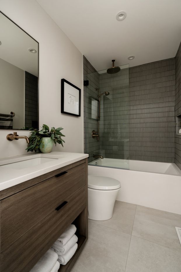 A compact contemporary bathroom with a wall-mounted dark wood vanity, white integrated basin, brushed bronze tapware, toilet, and combined bathtub-shower. The shower walls are clad in narrow vertically patterned grey-green tiles, while the floor uses large light grey tiles. A large frameless mirror, recessed ceiling lights, framed artwork, greenery, and folded towels complete the refined space.