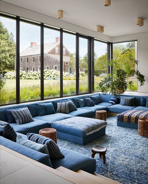 A bright, spacious living room with a large deep-blue sectional sofa arranged around a matching upholstered ottoman, striped blue cushions, woven side tables, and a pale blue textured rug. Floor-to-ceiling black-framed glazing wraps around the room, overlooking a landscaped garden and a traditional shingle-clad house. Potted greenery adds an organic element, while simple cylindrical pendant lights and a clean white ceiling create a refined contemporary setting.