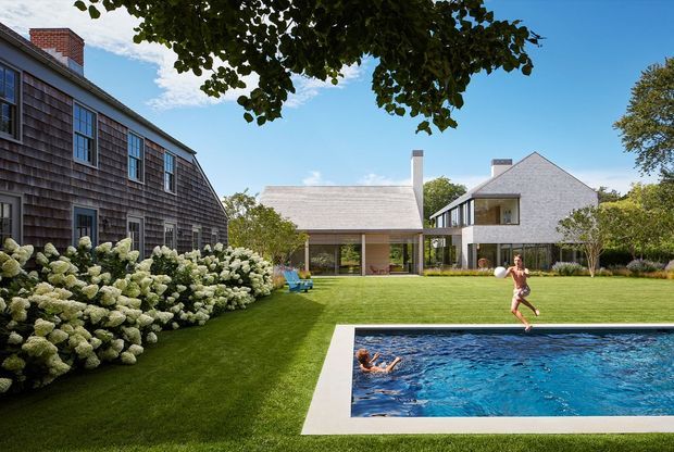 A landscaped residential property viewed from the garden, featuring a rectangular swimming pool, expansive lawn, mature planting, and several connected buildings. The composition includes a weathered shingle-clad structure at left, a low central pavilion with a pitched roof, and a contemporary pale-clad two-storey house at right.