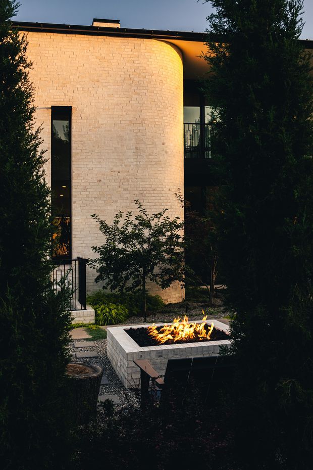 Dusk exterior view of a contemporary residence with a curved pale brick facade, narrow dark-framed windows, and a flat roof. Dense planting frames a landscaped courtyard containing a square raised brick outdoor fire pit with visible flames, gravel paths, and dark metal railings. Warm architectural lighting highlights the curved wall and creates a dramatic, intimate atmosphere.