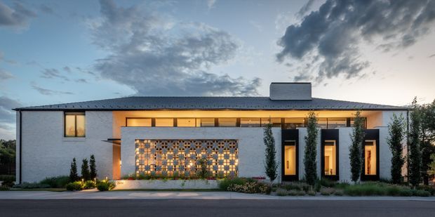 A low, single-storey contemporary residence viewed from the street at dusk. The long white rendered facade sits beneath a shallow dark pitched roof with a central rectangular chimney or roof element. Warmly illuminated timber-toned openings and tall glazed entry bays are screened by slender cypress-like planting. A prominent decorative breeze-block or perforated masonry screen forms a patterned feature wall near the entrance, with landscaped planting along the frontage.