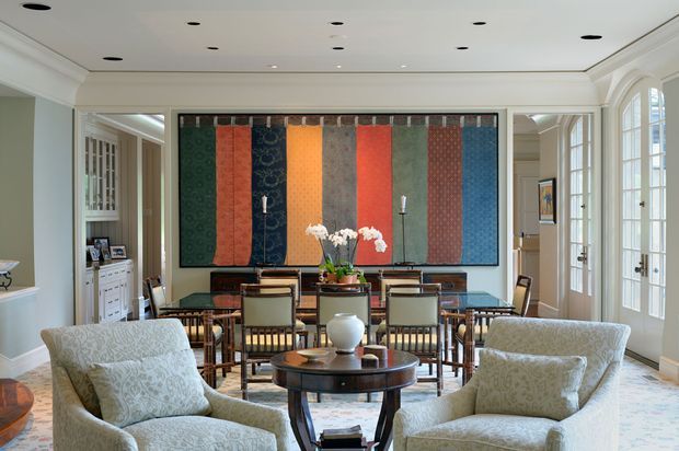 Elegant formal sitting and dining room with a large colourful abstract textile artwork, paired upholstered lounge chairs in the foreground, and a dark wood dining table with upholstered chairs behind. The room features a coffered-style white ceiling with recessed lighting, pale walls, ornate white trim, tall French doors, built-in cabinetry, and a patterned light rug. The overall look is refined and traditional with a curated contemporary art accent.