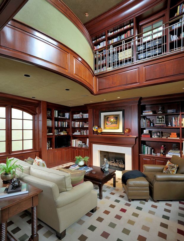 A richly detailed traditional library-style living room with extensive dark timber wall panelling, built-in bookshelves, a central fireplace with mantel, and a mezzanine-level gallery overlooking the room. Upholstered sofas and leather seating surround a dark timber coffee table. The room features arched window and ceiling details, decorative railing, framed artwork, books, and a multicoloured geometric area rug. The overall atmosphere is warm, refined, and scholarly.