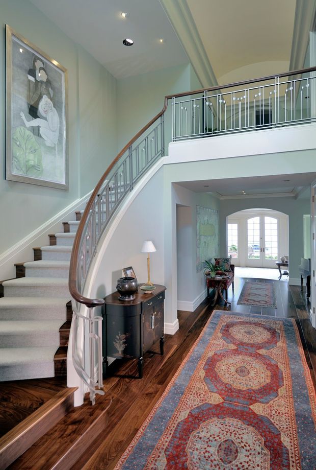Elegant residential entry hall with a sweeping curved staircase, light carpeted treads, dark timber flooring, decorative metal balustrades with a timber handrail, and a mezzanine-level landing. Pale blue-green walls, traditional trim, patterned runners, artwork, and a small console create a refined, formal interior. An arched opening leads toward a bright room with glazed doors at the rear.