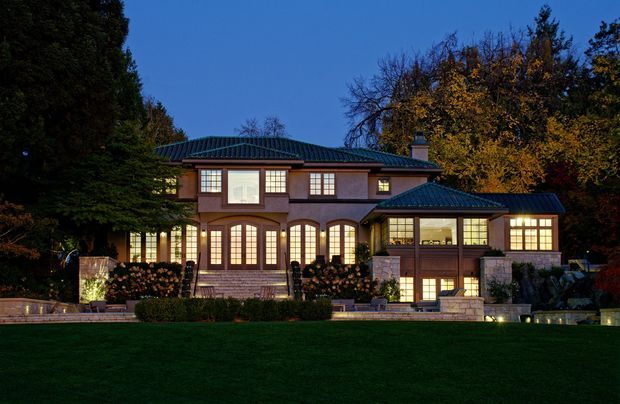 A large, symmetrical two-storey residence photographed at dusk, set behind a broad manicured lawn and mature trees. The house has a dark hipped metal roof, warm-toned masonry or rendered walls, extensive multi-pane windows and French doors, projecting upper-level bays, stone-clad lower walls, and carefully arranged exterior lighting. A lower side wing and landscaped garden elements are also visible.