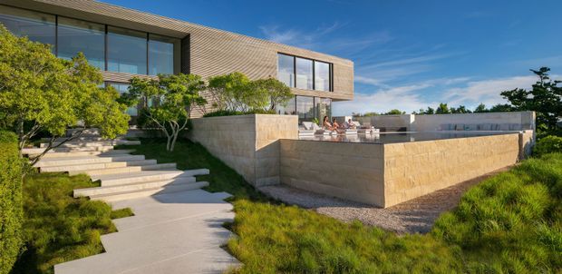 Modern two-storey residence with extensive floor-to-ceiling glazing, horizontal timber-toned cladding, and large sandstone or rendered masonry retaining walls. Broad concrete steps and a winding path lead through landscaped grasses and small trees to an elevated outdoor terrace with seating. The composition emphasizes the contemporary building form, outdoor living platform, and landscaped approach under clear blue skies.